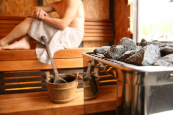 A person in a towel sits on a bench in a wooden sauna, the stove visible in the foreground.