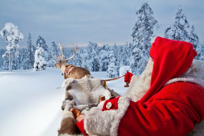 A reindeer pulls Santa Claus in a sleigh in a snowy forest landscape.