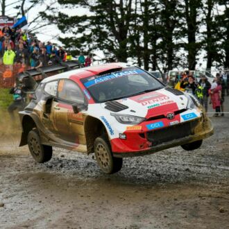 A racing car is about half a metre in the air above a muddy road as fans watch in the background.
