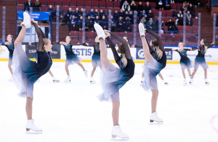 A dozen girls in matching outfits skate across an ice rink, some of them on one leg while holding the other leg above their heads.
