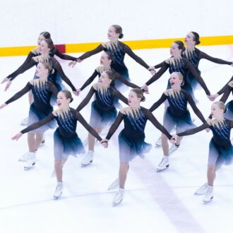A group of girls in matching dresses is skating across an ice rink in coordinated lines of four skaters holding each others' hands.