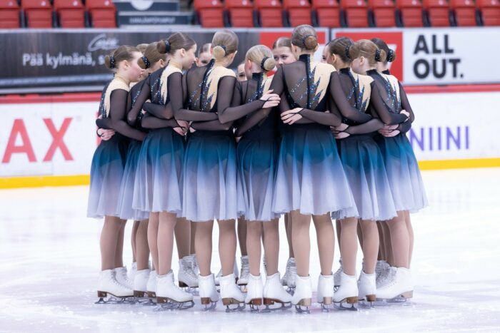A group of girls in matching outfits stand on an ice rink in a circle with their arms around each other.