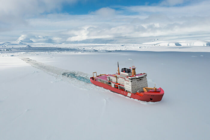 A large red ship is clearing a path across a bay covered in ice and snow, with glaciers in the background.