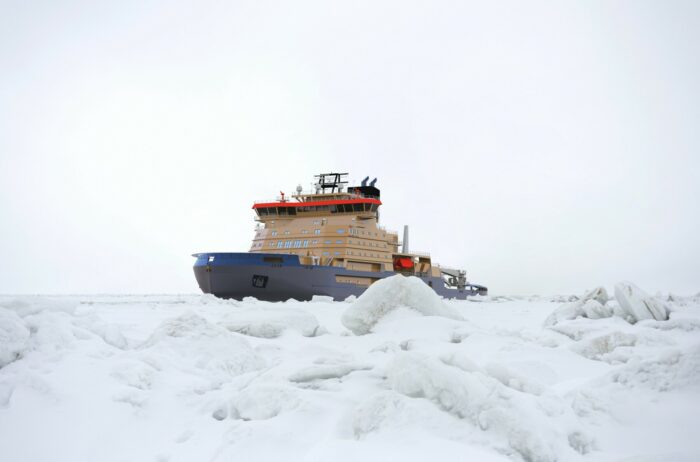 A large ship is progressing through a wintery sea, partially obscured by hunks of ice piled in the foreground.