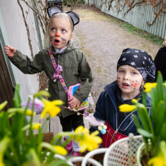 Several children dressed in costumes are at the door of a house beside flower pots full of daffodils.