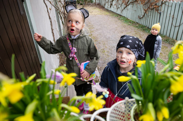Several children dressed in costumes are at the door of a house beside flower pots full of daffodils.