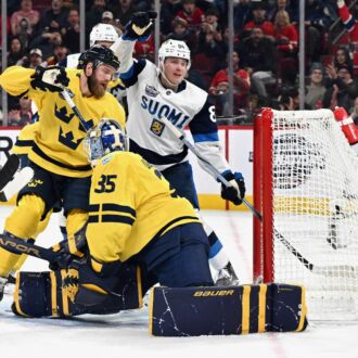 A Finnish ice hockey player raises his arm in triumph Finland scores a goal, while the Swedish goalie and a defender look forlorn.