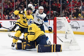 A Finnish ice hockey player raises his arm in triumph Finland scores a goal, while the Swedish goalie and a defender look forlorn.