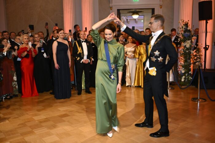 In a ballroom, a man and woman dance in front of spectators, everyone dressed in fancy formalwear.