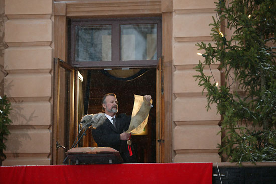 A man standing in a window, reading from an old-looking scroll.