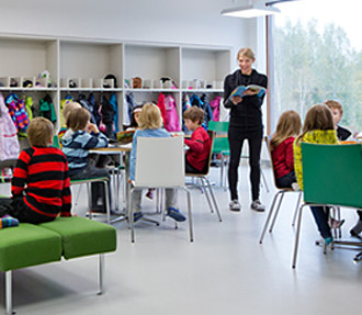 Schoolchildren in a light open space listening to their teacher reading a book.