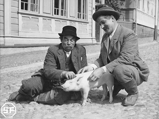 A black-and-white image of two gentlemen sitting in a cobblestone street petting a piglet. 