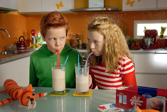 A red-haired boy and a blonde girl drinking milkshakes in a colourful kitchen.