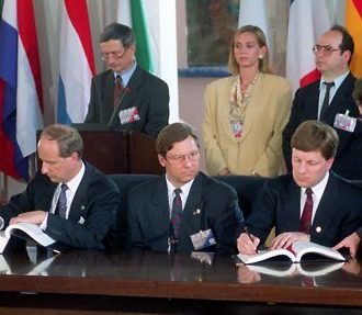 Four serious-looking people sitting at a table signing papers, while six other people are standing behind them.