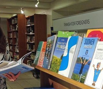 A smiling dark-haired woman browsing Finnish textbooks at a bookstore.