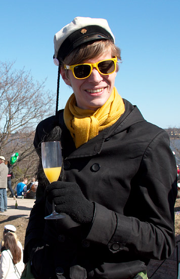 A smiling student in yellow sunglasses and scarf and the graduation cap holding a glass of Mimosa.