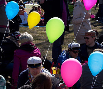 A large crowd of people picknicking at a park with colourful balloons.