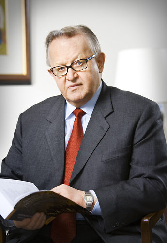 A main in a suit, tie, and glasses, with an open book in his hands, looks at the camera.