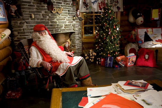 Santa Claus sitting in a rocking chair by a fireplace; a Christmas tree in the background.