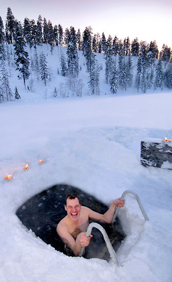 A widely smiling man dipping in a hole in the ice.