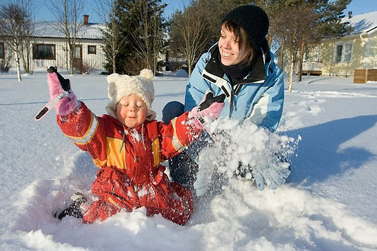 A small child dressed in coveralls sitting in the snow, throwing the snow upwards; an older person next to the child.