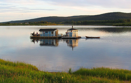 Two small sauna buildings are floating in a calm river. A boat is tied to one of the buildings. There are islands and forested hills in the background. 