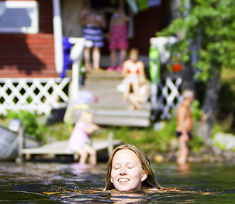 Two people are swimming in a lake. In the background, there is a dock and a sauna building with people sitting on the porch.
