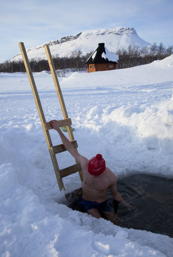 Um homem vestindo um gorro vermelho e calção de banho preto está descendo uma escada em um buraco em um lago congelado. Ao fundo há uma pequena cabana e uma montanha nevada.