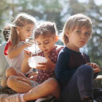 Varios niños de cinco o seis años posan en un bosque, sentados sobre una roca, uno de ellos comiendo de un recipiente de plástico.