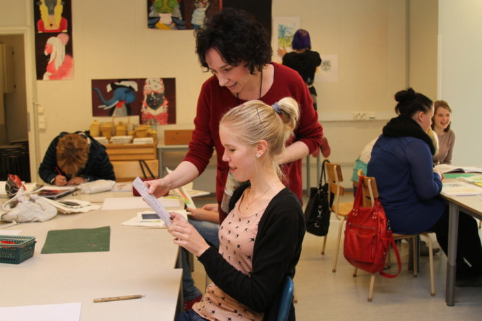 Teacher checking a students paper; other students studying in the background.