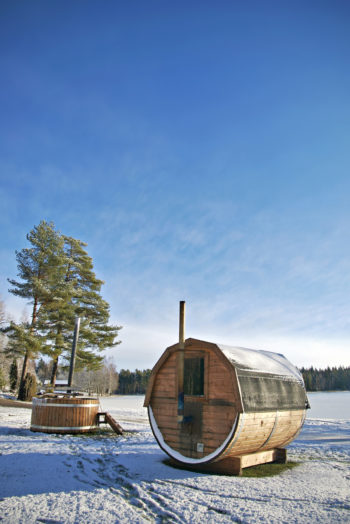 雪景中，冰封的湖边有一座桶状的桑拿屋和一个热水浴缸。