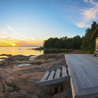 There is a cabin with a large terrace on a beach of smooth rocks. The sun is setting into the sea in the background.