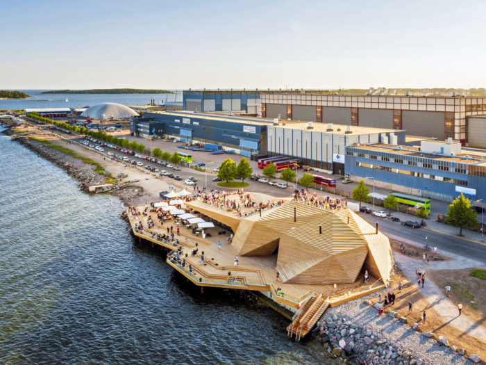 Масса людей на террасе современного деревянного здания геометрических форм у моря, на заднем плане несколько промышленных зданий. 