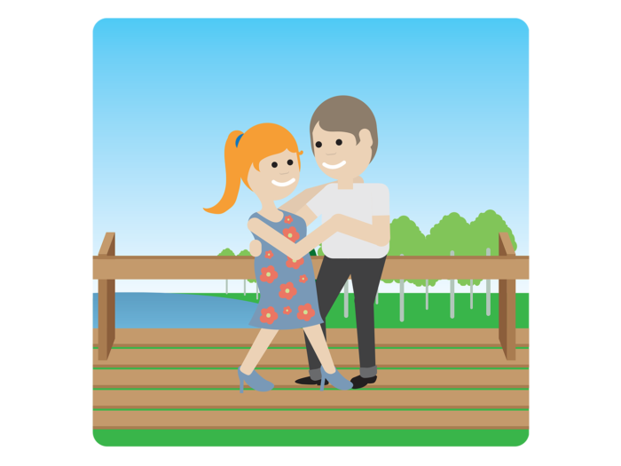 A smiling woman and man are dancing on an outdoor wooden platform, with a lake and some trees in the background.