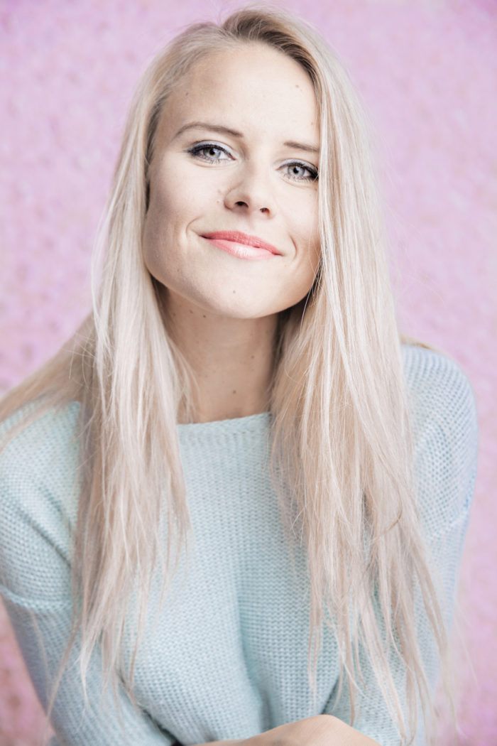 Portrait of a smiling Nelli Lähteenmäki on a pink background.