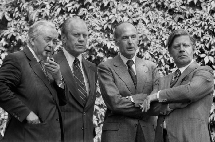 A black-and-white photo of Harold Wilson, Gerald Ford, Valery Giscard d’Estaing and Helmut Schmidt standing in front of a hedge.