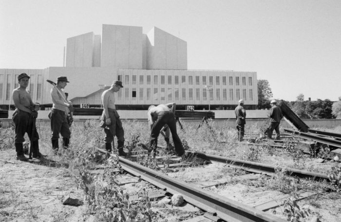 A black-and-white photo of shirtless young men digging the ground next to train tracks, Finlandia Hall in the background.