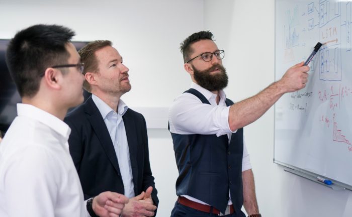 Three men looking intently at whiteboard with markings on it; one of the men pointing at something on the board.