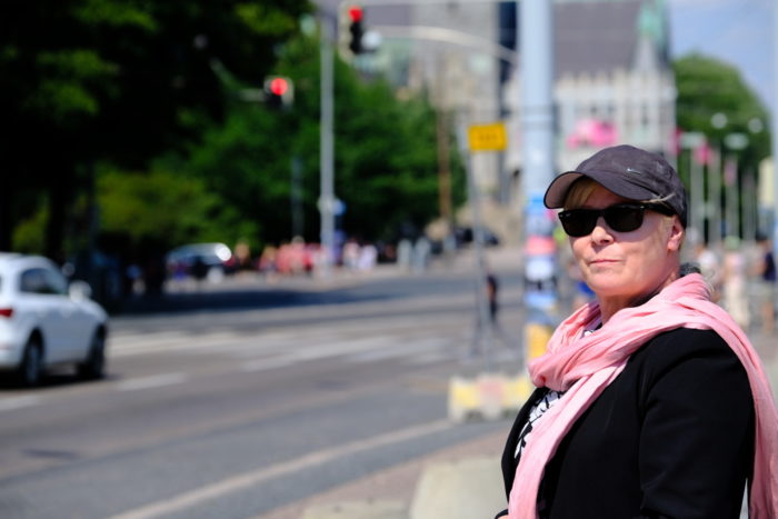 A woman in sunglasses, cap and a pink scarf standing by a road.