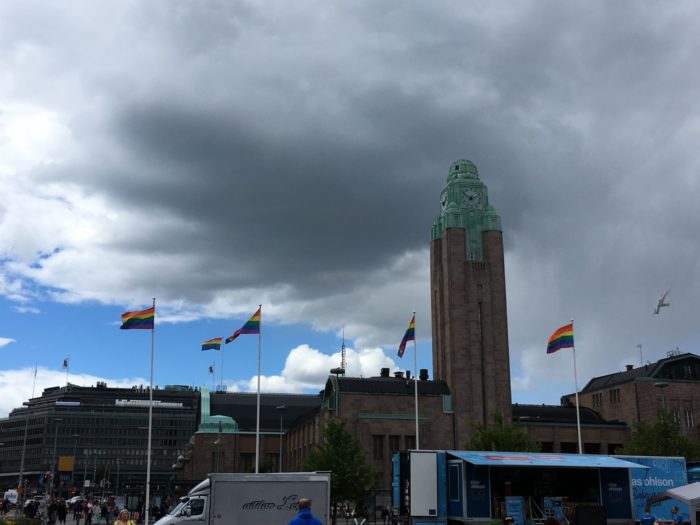 Several flagpoles have rainbow flags on them, in front of the Helsinki Railway Station's landmark tower.