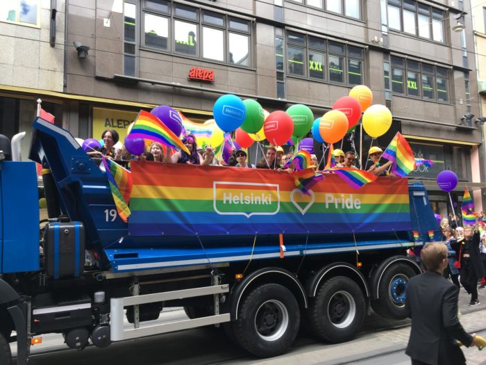 A rainbow-decorated truck full of people with balloons and with a sign that says Helsinki Pride.