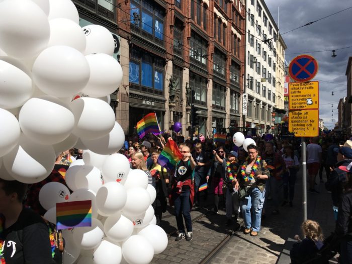 A colourful parade progresses down a main street in Helsinki, with a cloud in the sky far behind it.