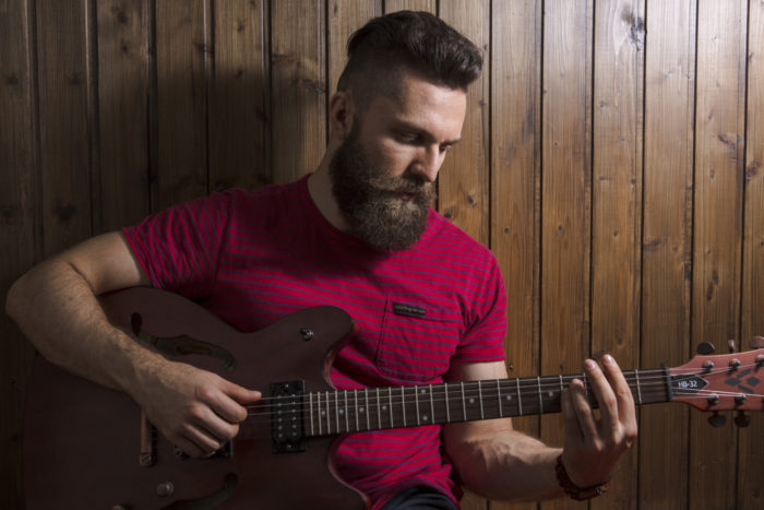 A bearded man playing the guitar in front of a wood paneled wall. 
