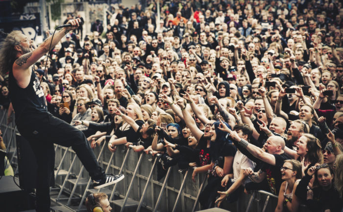 A musician on a stage plays guitar while a sea of fans waves their arms.