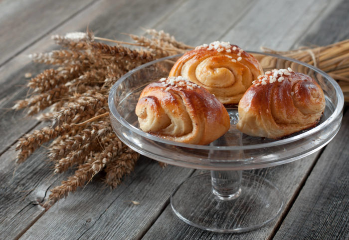 Three cinnamon buns on a glass tray on a wooden table, a bunch of wheat behind the tray.