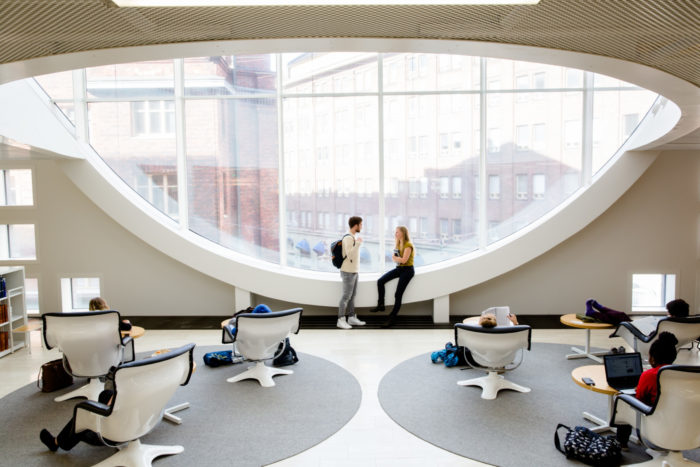 Two students hanging by a big oval-shaped window, other students studying in chairs nearby.