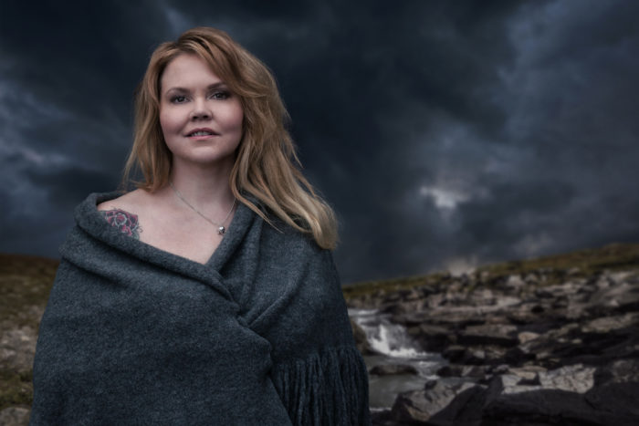 Portrait of author Katja Kettu standing outside in a rocky landscape and a stormy-looking sky.