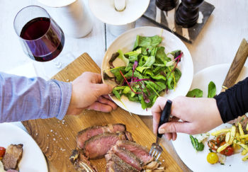 On a table there is a glass of red wine, a bowl of salad and a cutting board with sliced beef; hands from two different people are in the process of picking up food.