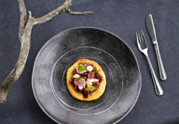 A fancy plate of food sits next to a tree branch on a grey tablecloth.