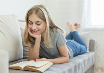 A smiling girl lies on a couch reading a book.